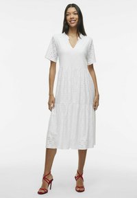 Robe en coton blanche avec jupe à volants, manches courtes et décolleté en V ornée de broderies en œillets. Assortie avec des sandales à talons hauts à lanières rouges.