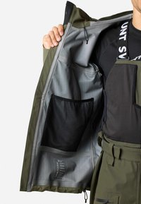 Grüne wasserdichte Jacke mit grauem Innenfutter, ausgestattet mit einer Netzinnentasche und Beschriftung. Das Design umfasst einen Frontreißverschluss und verstellbare Bündchen.