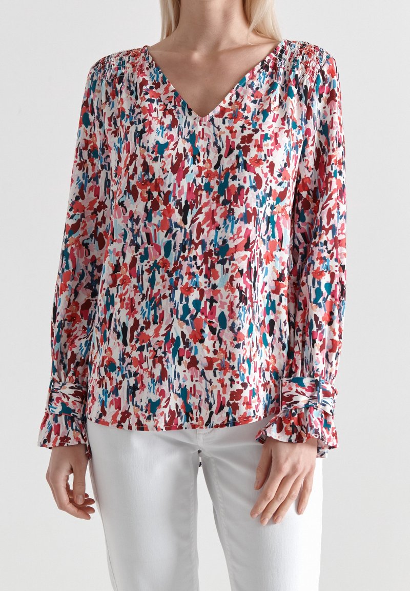 Blusa floreale astratta con scollatura a V, maniche lunghe e dettagli di polsini arricciati. I colori includono rosa, rosso, blu e bianco su uno sfondo chiaro.