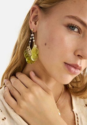 Mujer rubia con piel clara lleva pendientes colgantes con discos translúcidos verdes y cuentas blancas, sosteniendo el cuello con una mano.