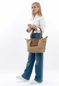 Borsa tote beige con dettagli in pelle marrone, doppie maniglie, tasca con zip, ampia internamente e realizzata in materiale morbido. Modello che indossa un abbigliamento casual.