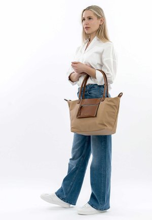 Beige tote bag with brown leather accents, dual handles, zippered pocket, spacious interior, and soft material. Model wearing casual outfit.