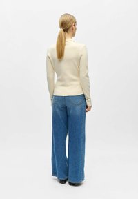 Femme avec queue de cheval blonde portant un haut crème à manches longues et un jean bleu à jambes larges, debout de dos sur un fond blanc uni.