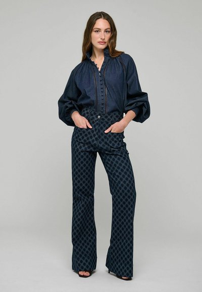Femme portant un chemisier bleu foncé froncé avec des détails de boutons et un pantalon large à motifs, debout les mains dans les poches devant un fond uni.