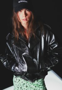 Černá kožená dvouřadová bunda s lesklou texturou a knoflíkovými detaily, spárovaná se zelenou květinovou sukní pokrytou flitry a černou čepicí.