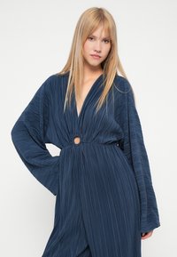 Mārīnas krāsas platas bikses ar dziļu V veida kaklu, platām piedurknēm un aplī veidotu izgriezumu jostas vietā. Mīksta, strukturēta auduma.