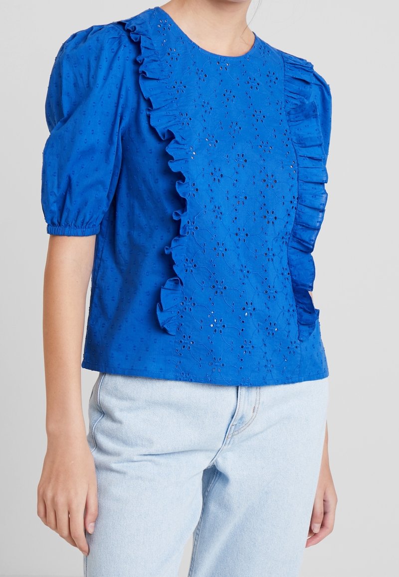 Blouse bleue à manches courtes avec motif floral ajouré et bordure à volants, portée avec un jean denim bleu clair sur une personne devant un fond gris.