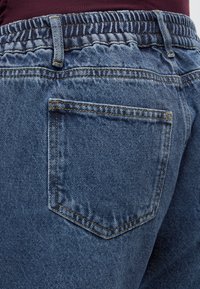 Nærbilde av blå denimjeans med elastisk midjebånd og synlig baklomme, brukt sammen med en burgunder topp.