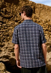 Uomo con camicia a maniche corte blu fantasia e occhiali da sole, in piedi di spalle alla fotocamera contro uno sfondo roccioso all'aperto.