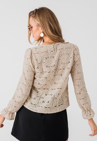 Beige bloemenkant blouse met lange pofmouwen, voorzien van een ronde halslijn en een zwarte rok. Het kant heeft ingewikkelde bloemenpatronen.