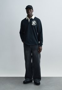 Μαύρο πόλο με μακριά μανίκια, εκρού γιακά και γραφικό αστροναύτη, συνδυασμένο με μαύρο τζιν φαρδιάς γραμμής και μοτίβο σε παπούτσια χωρίς κορδόνια.