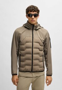 Fodrad jacka i beige med dragkedja fram, huva och svarta detaljer. Har ett sexkantigt kviltat mönster och muddar på ärmarna. Solglasögon på.