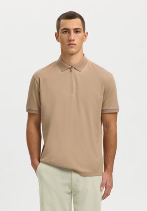 Tânăr cu păr scurt purtând un tricou polo bej cu mâneci scurte și fermoar în față și pantaloni deschiși la culoare, stând cu o mână în buzunar.