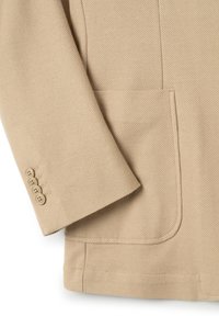 Giacca beige con texture, dotata di una tasca sul petto e polsini a quattro bottoni. Il materiale sembra tessuto e strutturato, aumentando la durata.