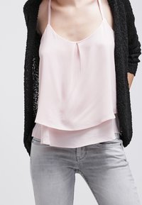 Haut en satin rose clair à couches avec de fines bretelles, présentant un détail plissé à l'avant, associé à un cardigan noir texturé et un jean en denim gris.