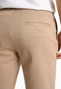 Pantaloni beige chiaro realizzati in tessuto liscio, dotati di una tasca posteriore con un unico bottone e una vita senza cuciture.