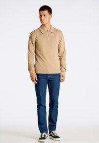 Beige stickad tröja med dragkedja i halsen, tillsammans med blå jeans och svarta sneakers, med en enkel design och enhetliga färger.