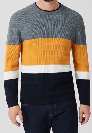 Homme portant un pull en maille côtelée avec des blocs de couleur horizontaux gris, jaune moutarde, blanc et bleu marine, associé à un jean foncé.