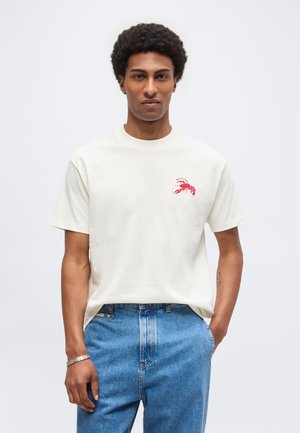 Hombre con pelo rizado que lleva una camiseta blanca con un gráfico rojo de langosta y vaqueros azules, de pie frente a un fondo claro liso.