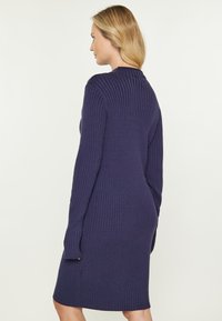 Gestreiftes Strickkleid in tiefem Marineblau. Es hat lange Ärmel und eine figurbetonte Silhouette, die oberhalb des Knies mit einem abgerundeten Saum endet.