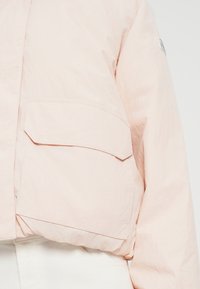 Veste courte rose clair en tissu texturé, avec une grande poche à rabat et des coutures ajustées.