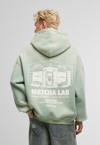 Sudadera verde claro de algodón, con una impresión gráfica en la parte posterior que incluye texto y una ilustración de una tienda. Textura suave, corte relajado.