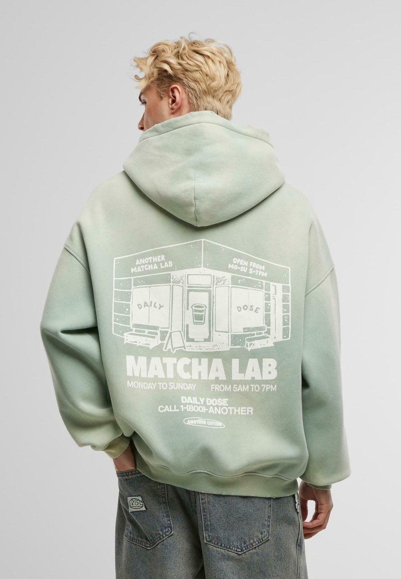 Sudadera verde claro de algodón, con una impresión gráfica en la parte posterior que incluye texto y una ilustración de una tienda. Textura suave, corte relajado.
