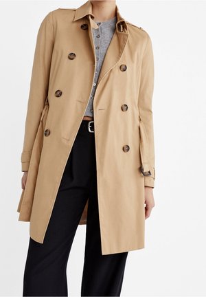 Femme portant un trench-coat beige croisé avec ceinture, par-dessus un haut gris boutonné et un pantalon noir, sur un fond uni.
