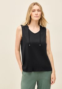Top nero senza maniche con scollatura a V e cordini al collo. Il tessuto sembra leggero, abbinato a pantaloni verde chiaro con texture.