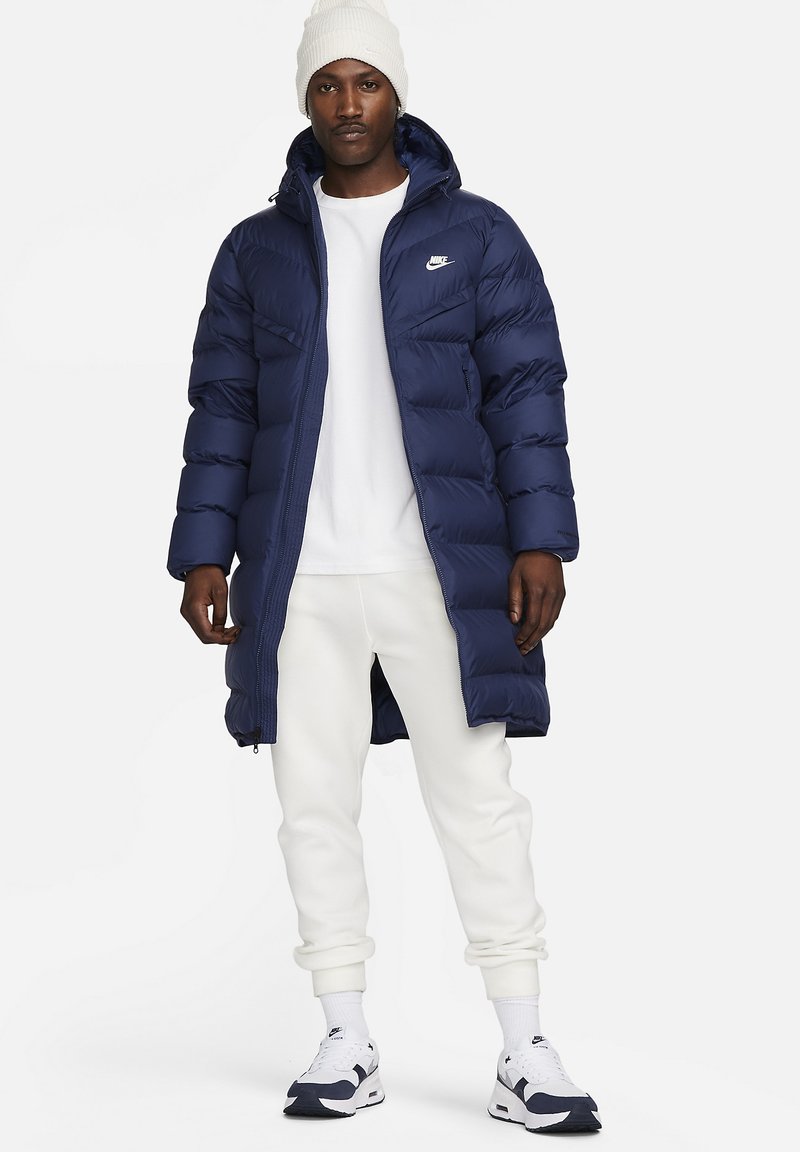 Marineblaue Steppjacke mit Kapuze, gestepptem Design, Reißverschluss und Logodetail auf der Brust, kombiniert mit weißen Hosen und Sneakers.
