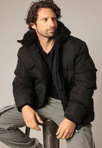 Doudoune noire avec col montant, fermeture éclair et design matelassé, portée sur une chemise noire. Pantalons cargo gris avec poches.