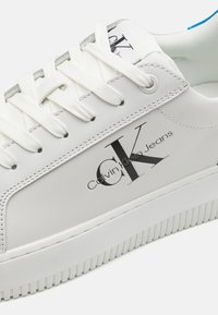 Bílé tenisky s hladkým koženým povrchem, s výrazným černým logem "CK" a textem "Calvin Klein Jeans". Hladká gumová podrážka.
