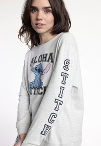 Mujer joven con cabello castaño que lleva puesta una sudadera gris clara con el personaje de dibujos animados Stitch en azul y el texto "ALOHA" y "STITCH" en la manga.