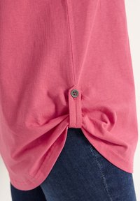 Vaaleanpunainen lyhythihainen paita, jossa on solmittu muotoilu sivussa ja napin yksityiskohta. Pehmeä rakenne ja rento istuvuus. Yhdistetty tummiin farkkuihin.