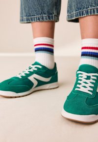 Baskets en daim vert avec accents blancs, dotées d'un design à lacets et d'une semelle texturée. Associées à des chaussettes blanches avec des rayures rouges, bleues et grises.