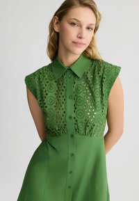 Abito verde con collo a camicia, maniche corte a cappuccio e pattern floreali ritagliati. Tessuto morbido con vita aderente e chiusura frontale a bottoni.