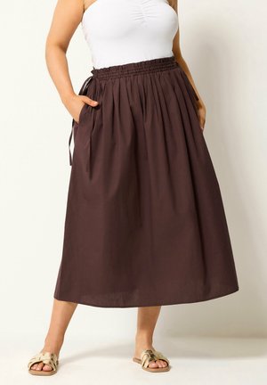Vrouw met een hoge taille, geplooide bruine midirok met zakken, gecombineerd met een witte mouwloze top en beige slides.