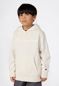 Chico con cabello lacio y negro, vistiendo una sudadera blanca de Champion con bolsillo frontal y logo en el pecho y la manga, de pie frente a un fondo liso.