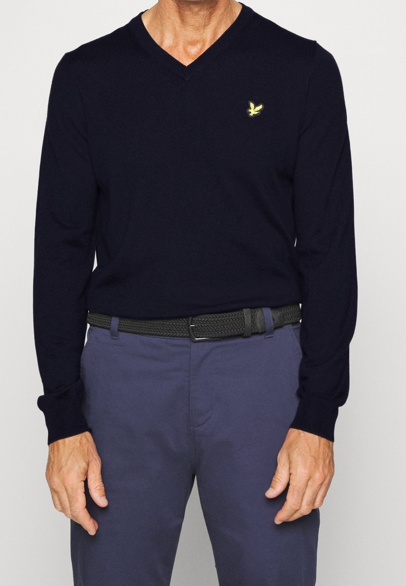 Pull en maille bleu marine à col en V en tissu doux avec manches longues, orné d'un petit logo jaune brodé sur la poitrine, associé à un pantalon bleu clair et une ceinture noire.