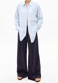 Personne portant une chemise à boutons rayée bleue clair ample et un pantalon large bleu marine, les mains dans les poches, se tenant sur un fond blanc.