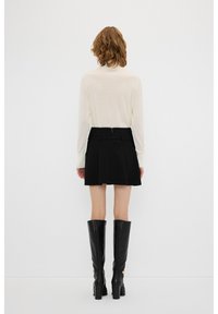 Schwarzer plissierter Rock mit einem taillierten Bund, kombiniert mit einem hellen Langarmshirt. Modell trägt knielange schwarze Stiefel.