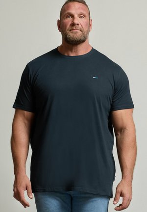 Muskelmand med skæg står med fronten fremad, iført en enkel mørk t-shirt og lyseblå jeans mod en neutral baggrund.