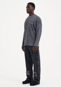 Maglietta grigia a maniche lunghe con righe e spacchi laterali, abbinata a jeans neri strappati con accenti grafici bianchi numerici e scarpe nere.