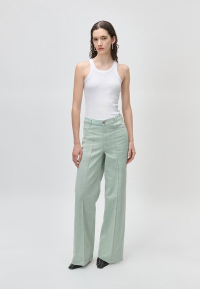 Γυναίκα με σγουρά μαλλιά που φοράει λευκό αμάνικο τοπ, ανοιχτοπράσινο παντελόνι με φαρδιά μπατζάκια και μαύρα μυτερά παπούτσια, στέκεται μπροστά σε ένα απλό φόντο.