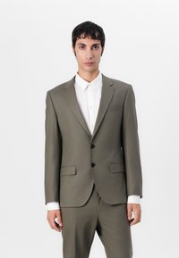 Olive green suit with notched lapels, two buttons, and side pockets. Paired with a white button-up shirt and visible cuff. Smooth texture.