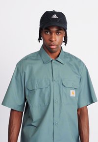 Smeđa, kratki rukavi, svijetlozeleni košulja na dugme s dva džepa na prsima i Carhartt logom. Nosi se s crnim šeširom s bijelim Adidas brandingom.