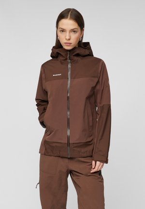 Donna con capelli castani lisci che indossa una giacca con cappuccio marrone e pantaloni abbinati, in piedi con le mani nelle tasche davanti a uno sfondo semplice.