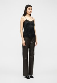 Γυναίκα που φοράει μαύρο καμήλο με δαντέλα, παντελόνι από σκούρο καφέ δέρμα και μαύρες μυτερές μπότες, στέκεται μπροστά σε απλό φόντο.