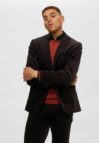 Svart kordfloret blazer med en enda ficka, bärs över en rostfärgad tröja. Modellen står med armarna i kors, vilket framhäver blazerns strukturerade design.