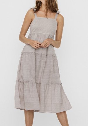 Šaty s čtvercovým výstřihem, vrstvené v světlé hnědé kostkované látce. Mají nastavitelné ramínka a volný siluetu s jemnými volány.
